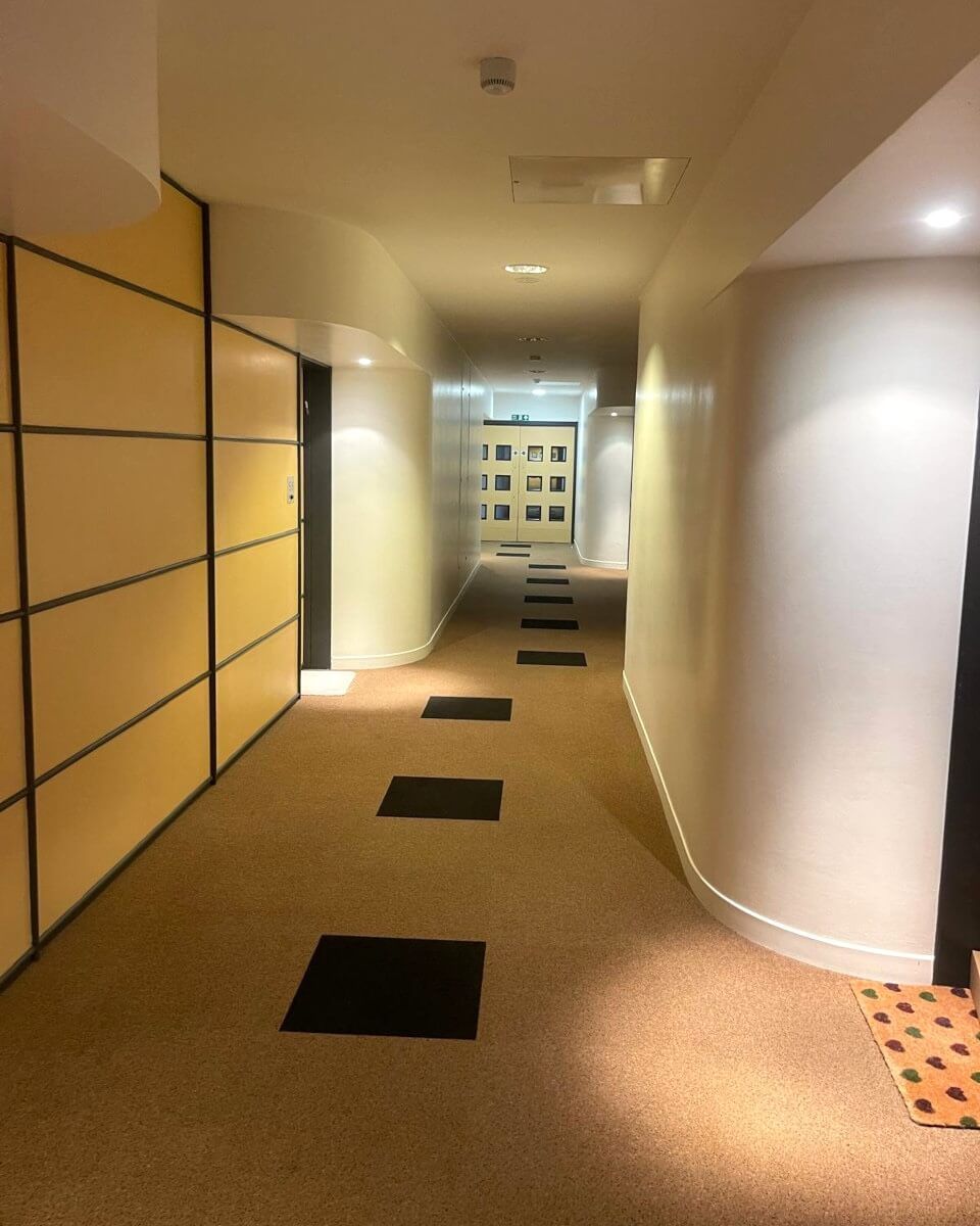 Long hallway with tan carpet and black square accents, leading to a door at the end. Yellow and white walls.