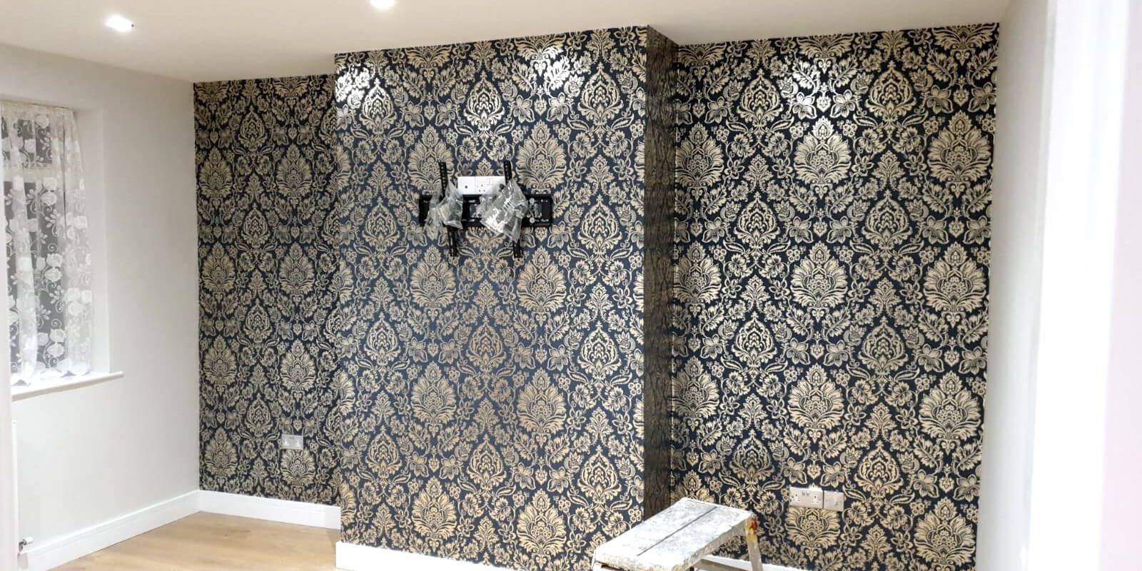 Room with ornate black and gold wallpaper, white trim, window with lace curtain, and wooden floor.