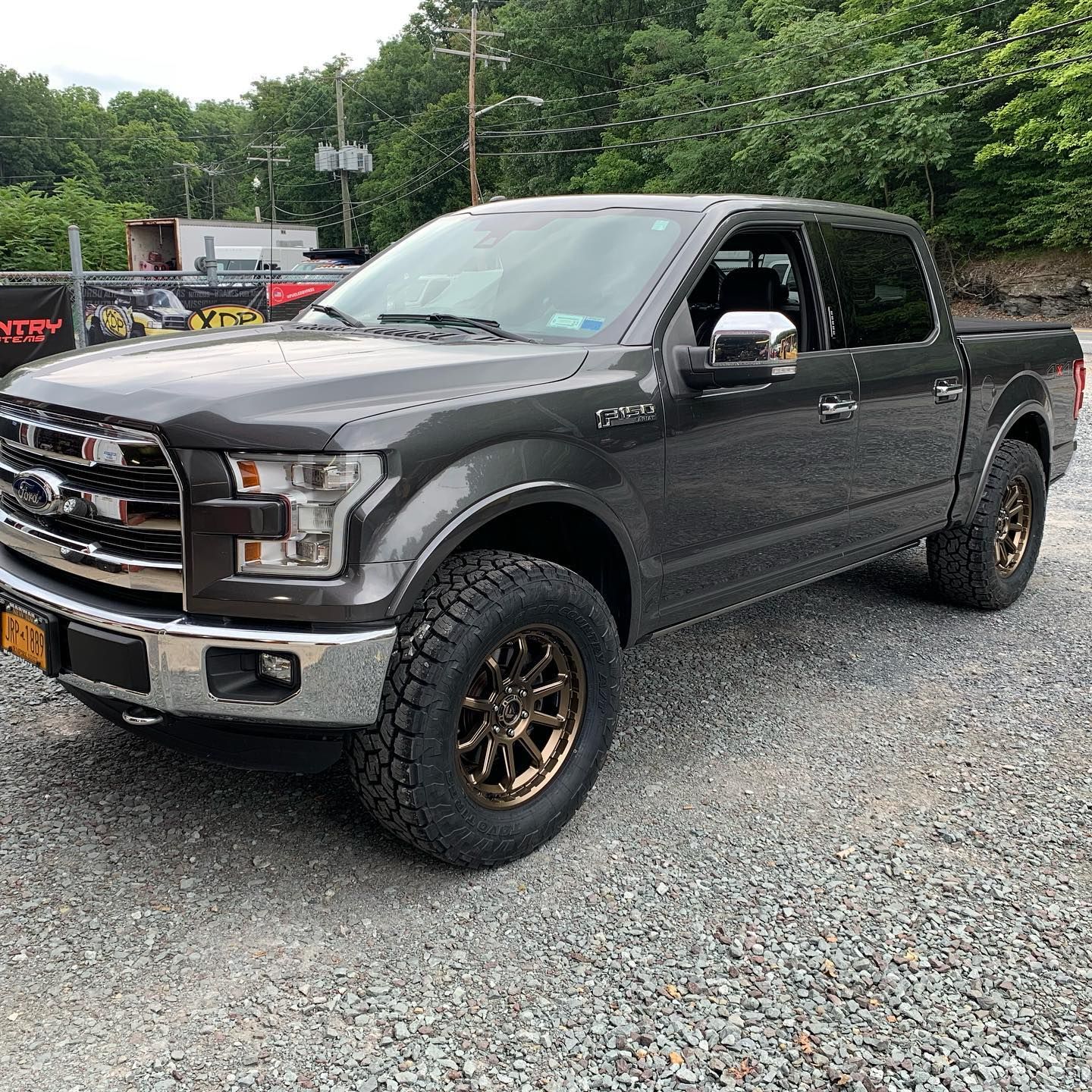 Gray Ford F-150 pickup truck with bronze wheels parked on gravel, in front of a building.