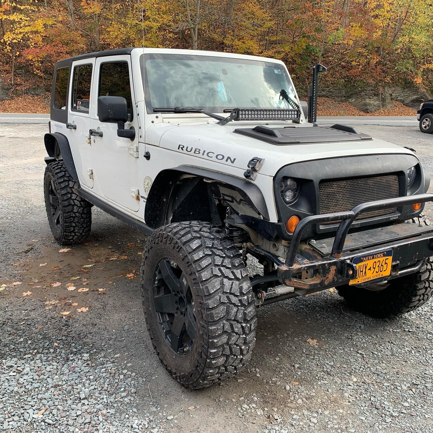 White Jeep Wrangler Rubicon with large tires, parked on a gravel surface in a wooded area.