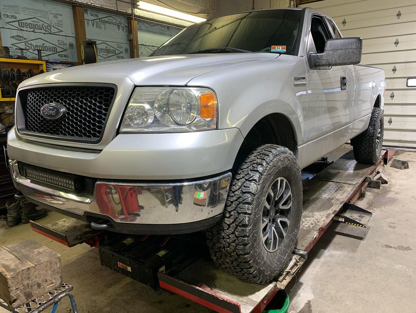 Silver Ford F-150 truck on a lift inside a workshop. Features black grille, off-road tires, and a light bar.