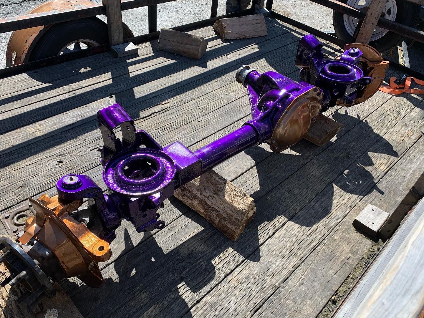 A bright purple car axle sits on wooden blocks on a trailer bed.