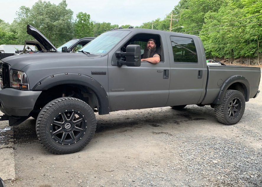 Man in gray pickup truck smiles at camera. Gray truck has black wheels and fender flares, parked outdoors.