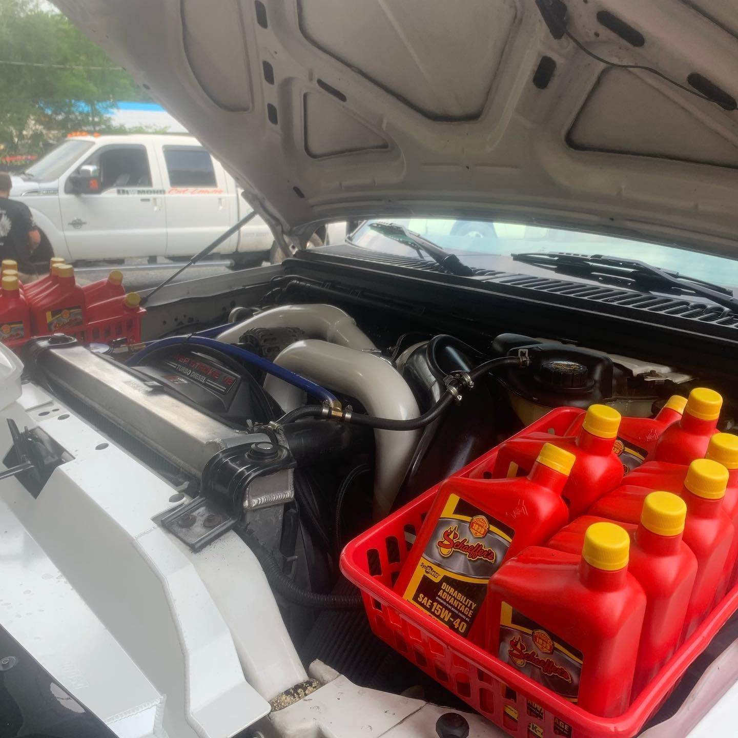 Open hood of a white truck. A basket of red oil bottles sits beside the engine. Another truck is in the background.