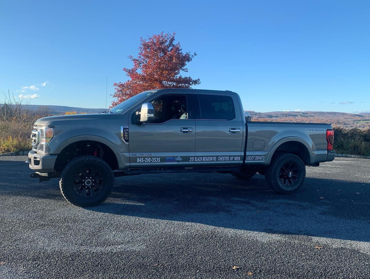 Gray Ford pickup truck with black wheels parked outdoors on a gravel lot; fall foliage in background. Gray Ford pickup truck with black wheels parked outdoors on a gravel lot; fall foliage in background.