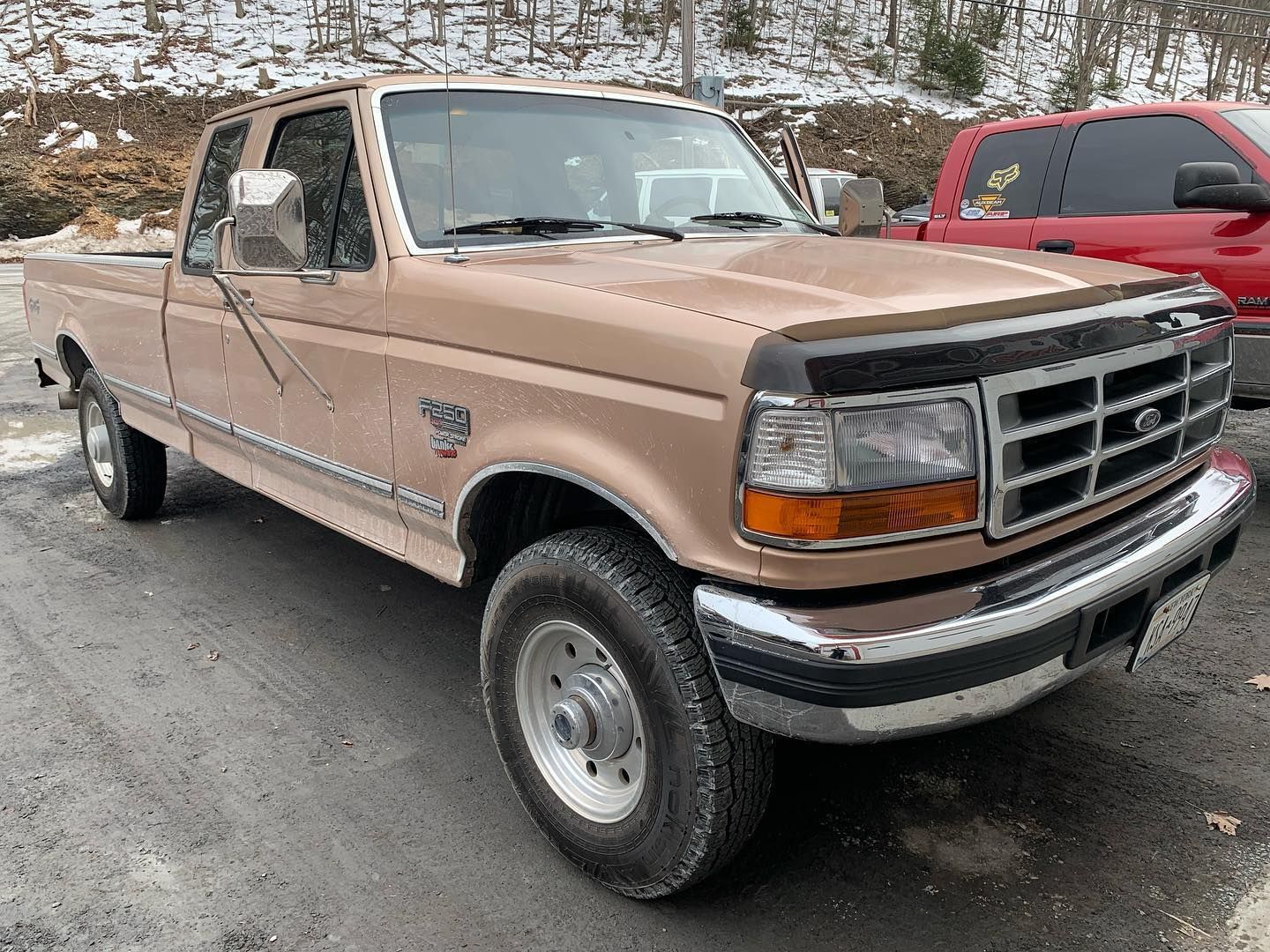 Tan Ford pickup truck parked on asphalt. Snowy background and red truck visible. Tan Ford pickup truck parked on asphalt. Snowy background and red truck visible.