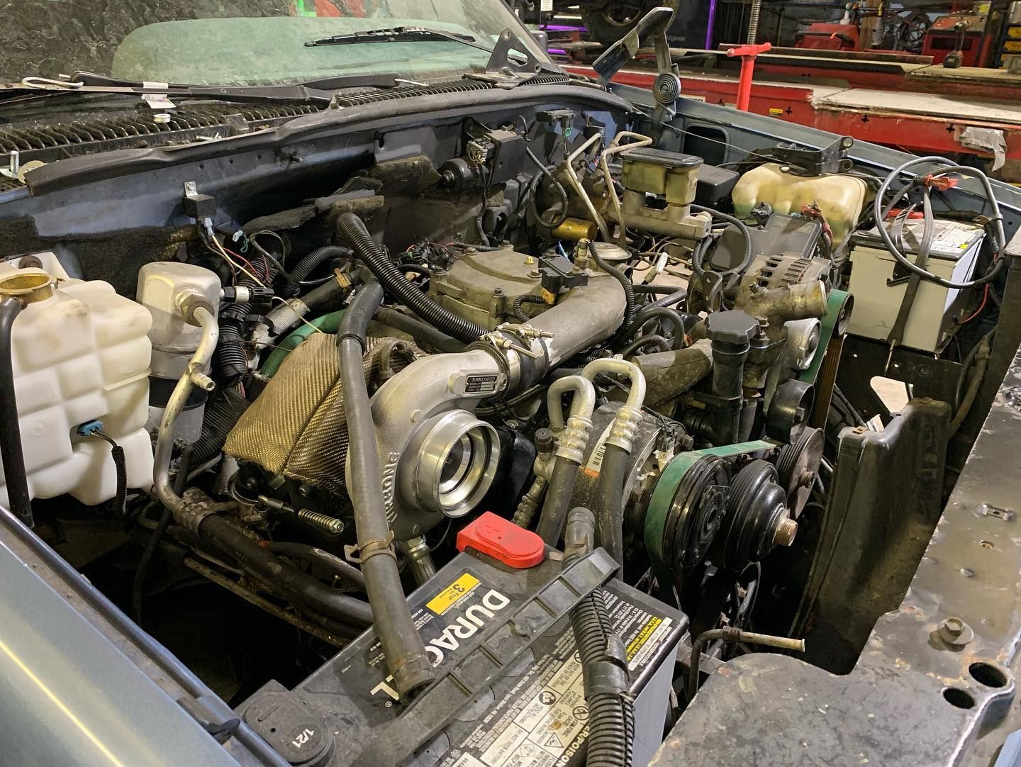 Engine bay of a vehicle with a large turbocharger and battery. Hood open, visible wiring and components.