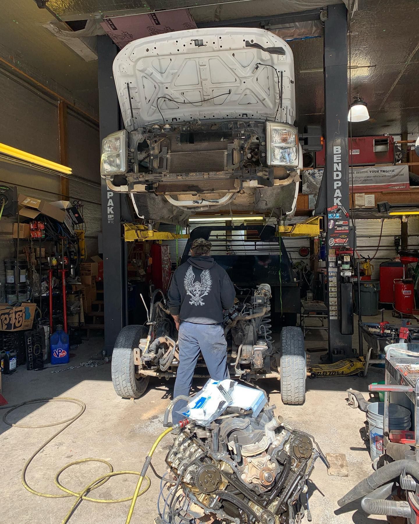 Truck being repaired in a shop, body on a lift, worker in front, engine on the ground.