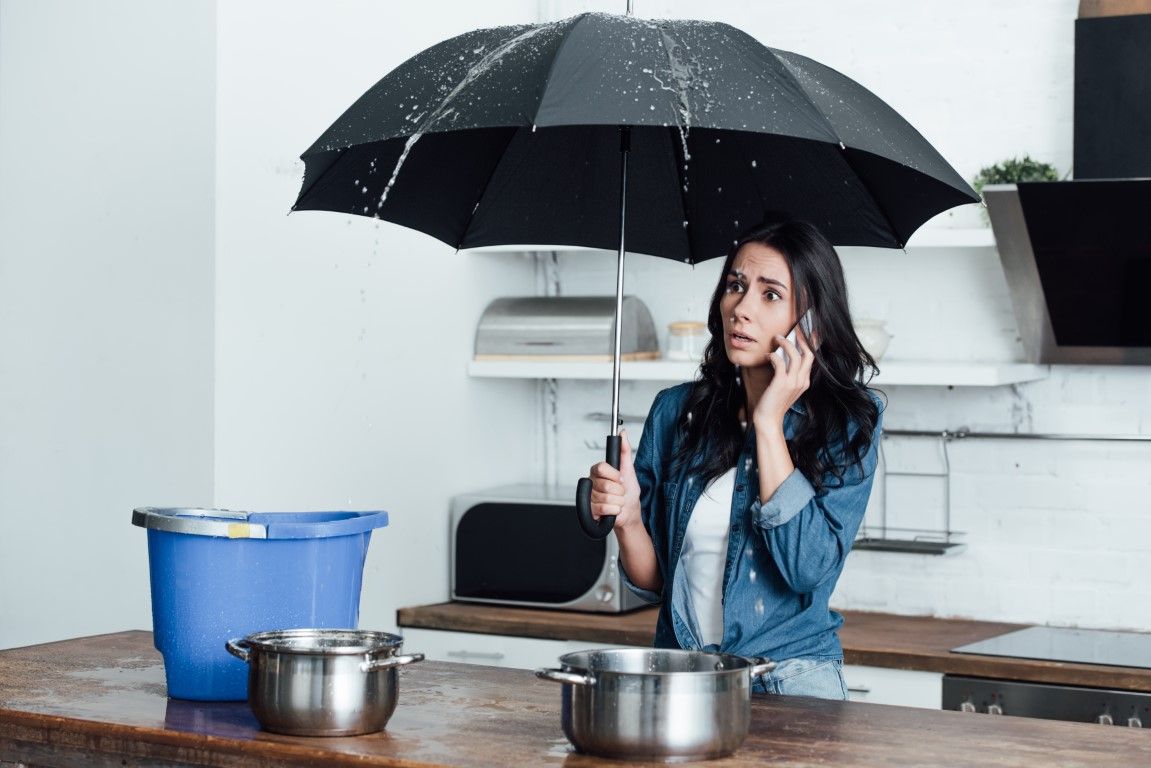 Woman in kitchen under an umbrella, talking on phone, looking concerned as water leaks from the ceiling. Buckets and pots collect water.