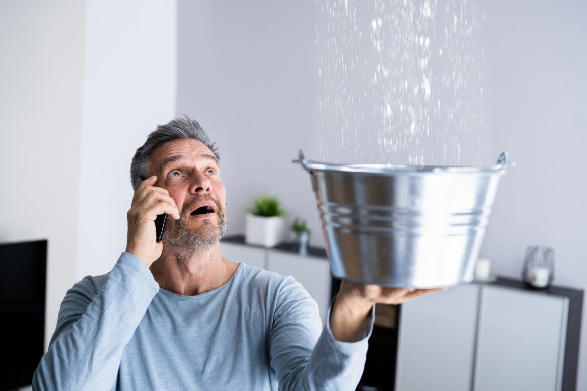 Man holding a bucket to catch water leaking from the ceiling, talking on a phone, looking worried.