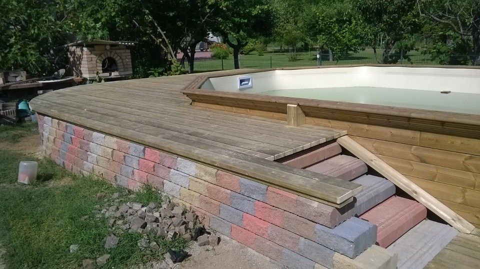 Terrazza in legno in costruzione accanto a una piscina fuori terra vuota, con assi impilate e un prato.