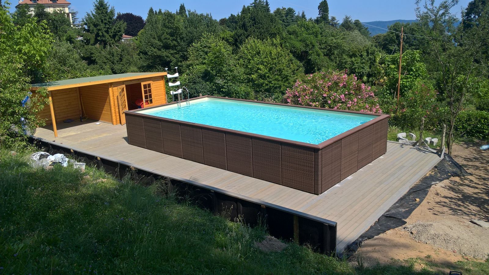 Terrazza in legno nel cortile sul retro con una piccola piscina rettangolare fuori terra e una casetta gialla accanto agli alberi.