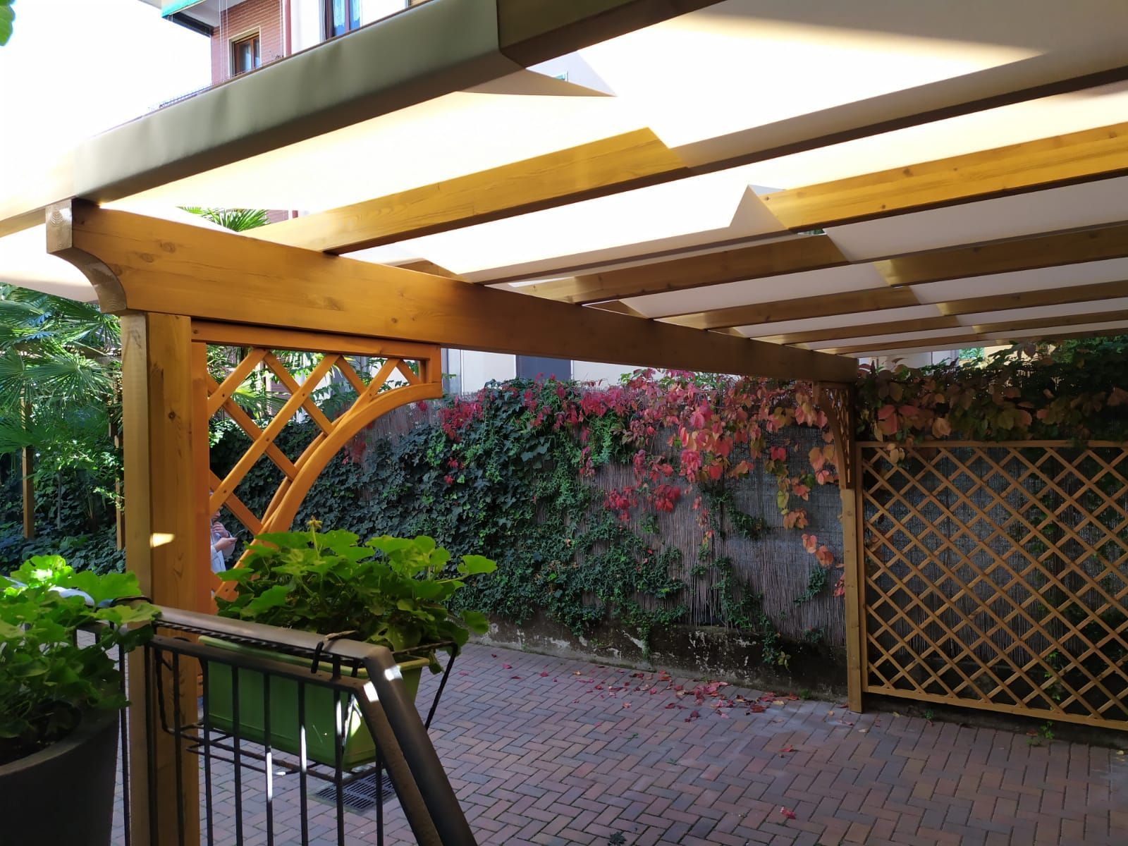 Patio coperto con travi in ​​legno, pergolato e cortile pavimentato in mattoni adiacente a un muro di cinta del giardino.