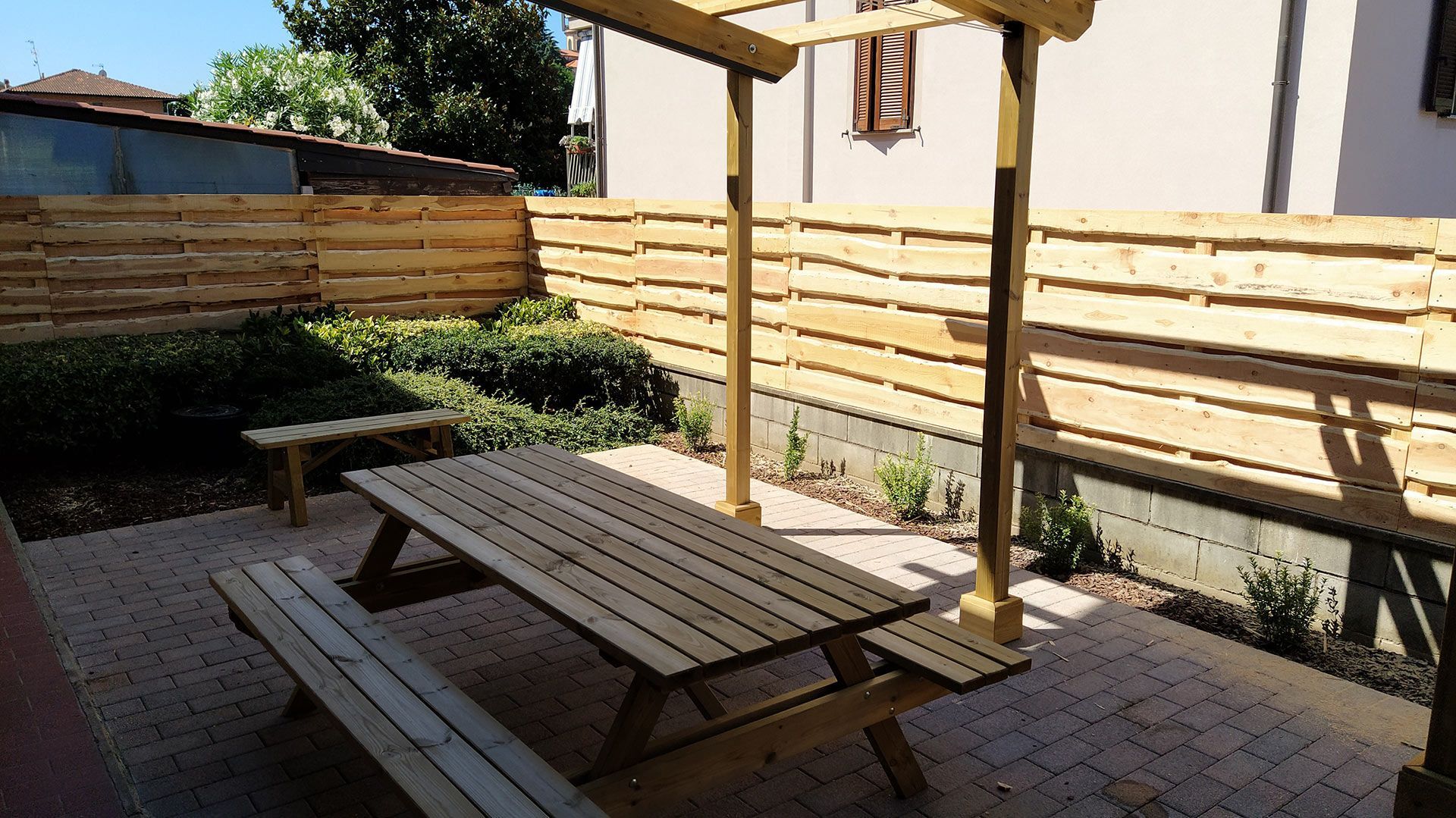 Patio esterno con tavolo da picnic in legno, portaombrelli, piante in vaso e recinzione in legno, esposto al sole.