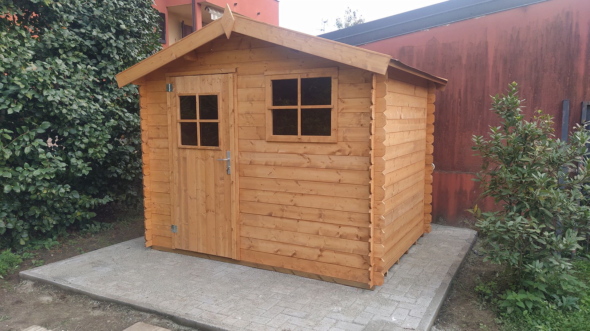 Casetta da giardino in legno con due finestre e una porta, posizionata su una lastra di cemento accanto a una recinzione rossa.