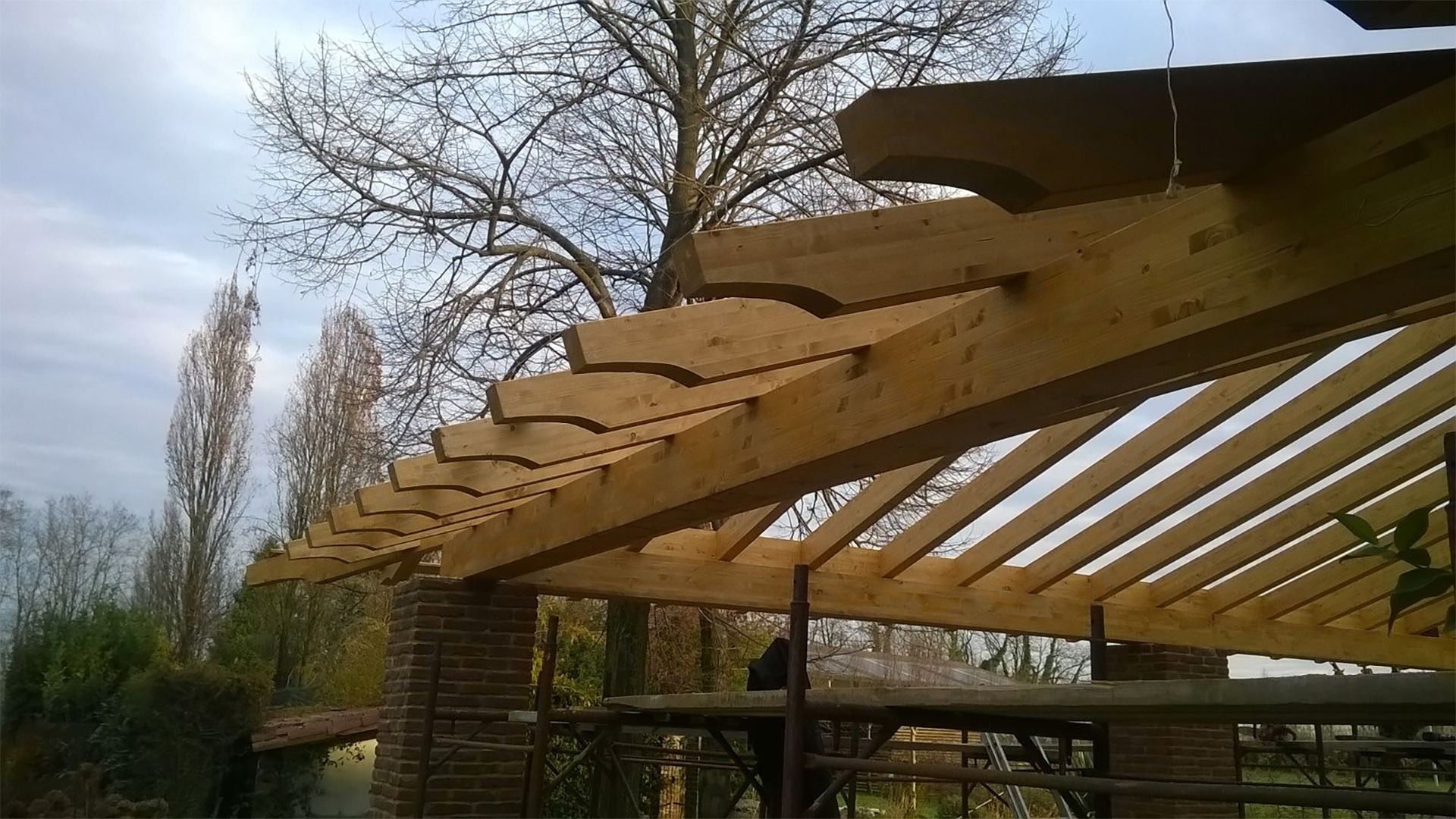 Travi di legno per il tetto di una pergola in fase di costruzione all'aperto, con alberi e cielo nuvoloso sullo sfondo.