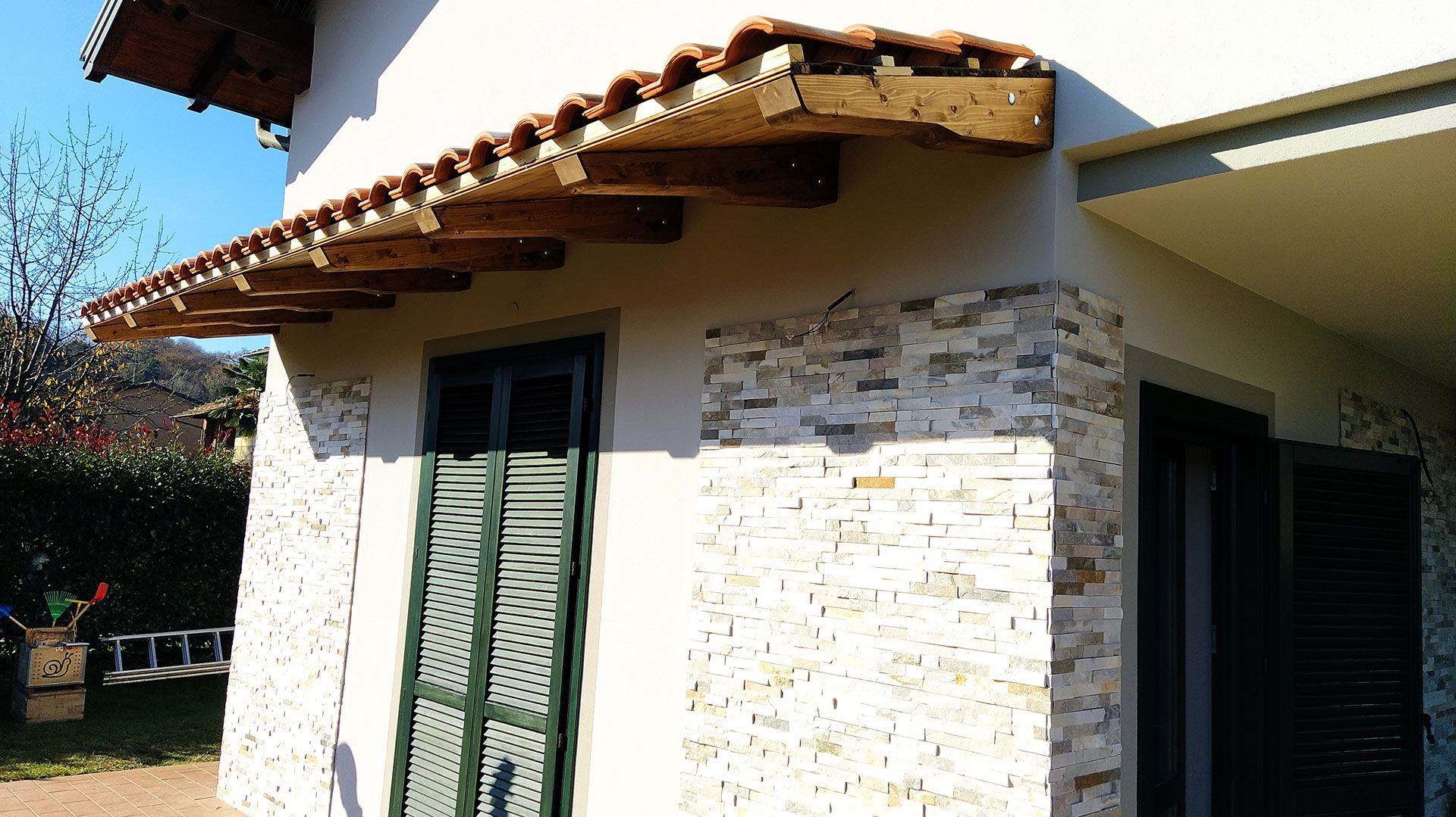 Piccola casa bianca intonacata con tetto di tegole, persiane verdi e muro di pietra alla luce del giorno