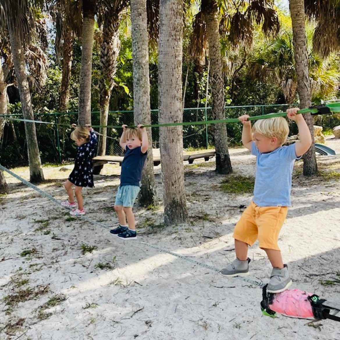 Three children are playing on a ropes course in the sand