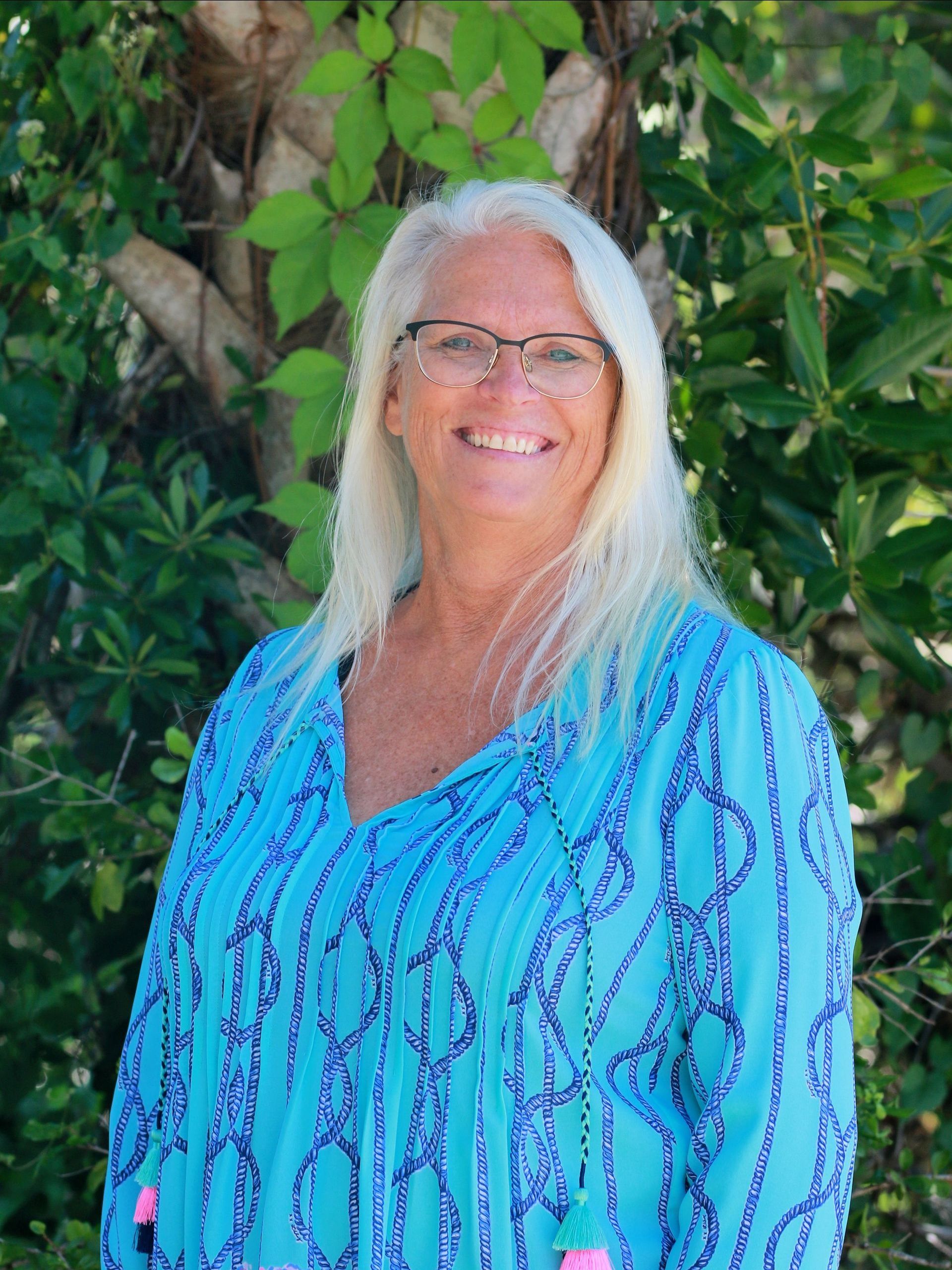 A woman wearing glasses and a blue shirt is standing in front of a tree.