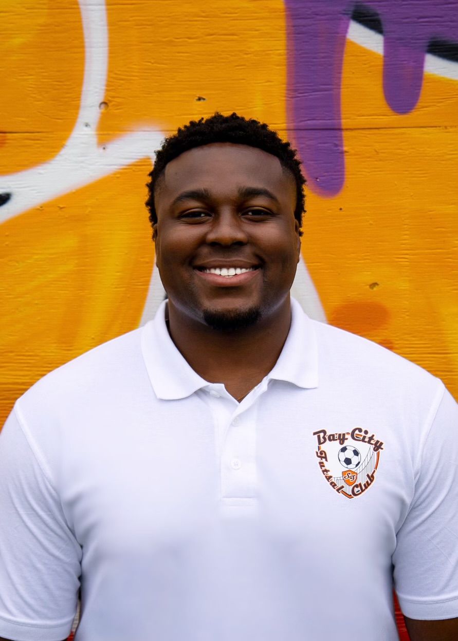 Ayinde Cole, Man in white polo shirt with logo, smiling in front of graffiti wall.