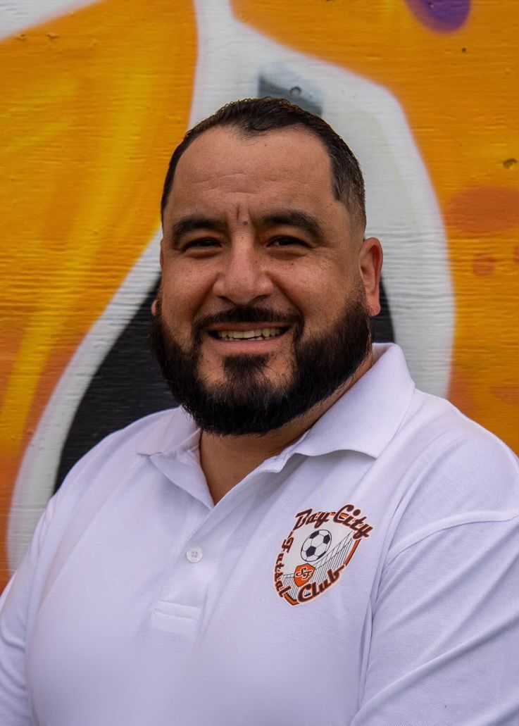 Antonio Medina , Man with beard, smiling, wearing white polo shirt with logo, in front of graffiti art.