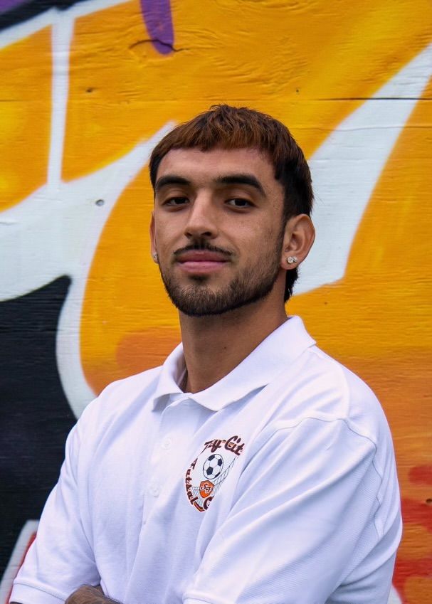 Adrian Medina, Man with short brown hair and beard, wearing a white polo shirt with logo, stands in front of graffiti.