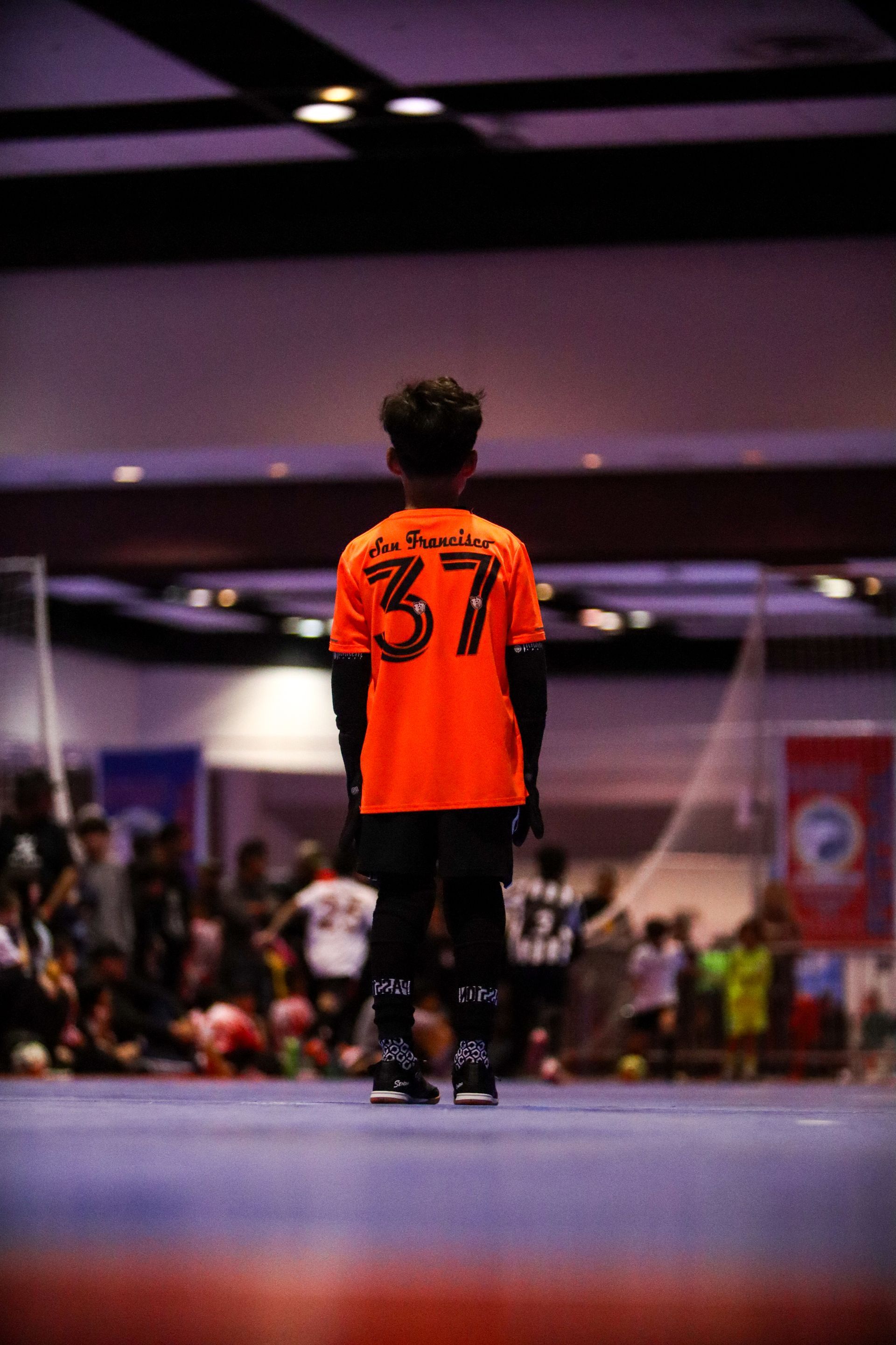 Boy in orange jersey, number 37, on indoor soccer field, facing away from camera, crowd in background. Bay city Futsal
