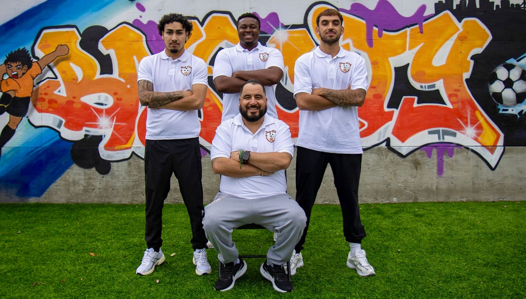 Bay city Futsal staff. Four people in white shirts and black pants pose in front of a colorful graffiti wall, with green grass below.