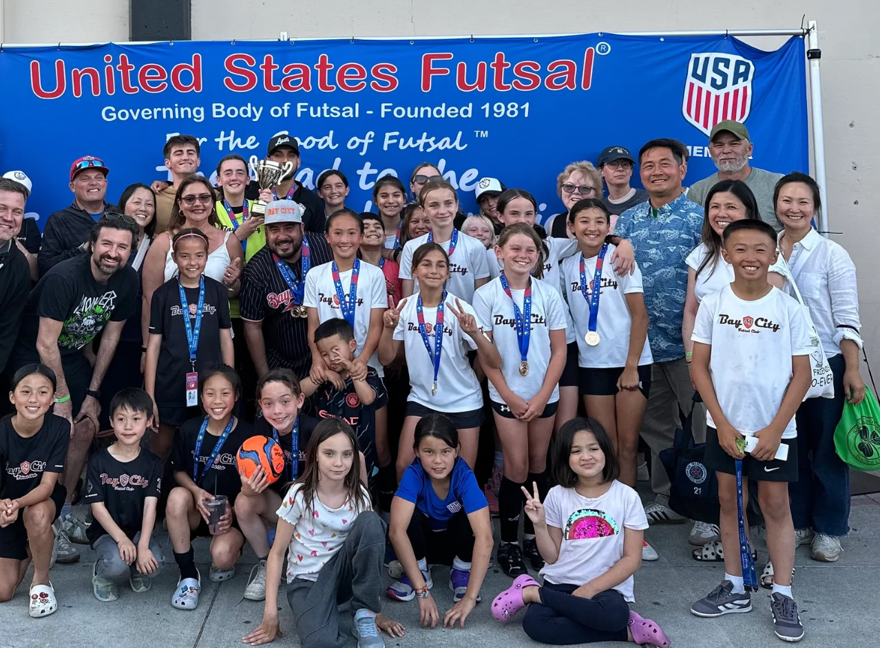 Group of people pose for photo in front of United States Futsal banner. Many wear medals; outdoor setting. Bay city Futsal