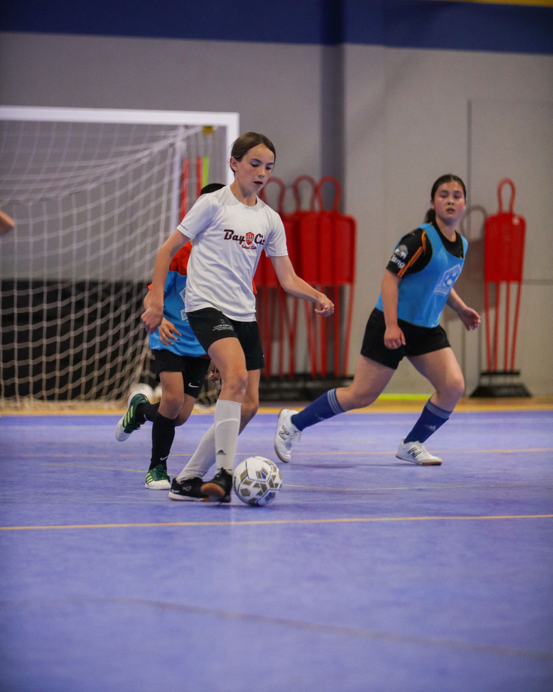 Youth soccer players on an indoor court. One dribbles the ball, two others chase. Blue and red color scheme. Bay city Futsal