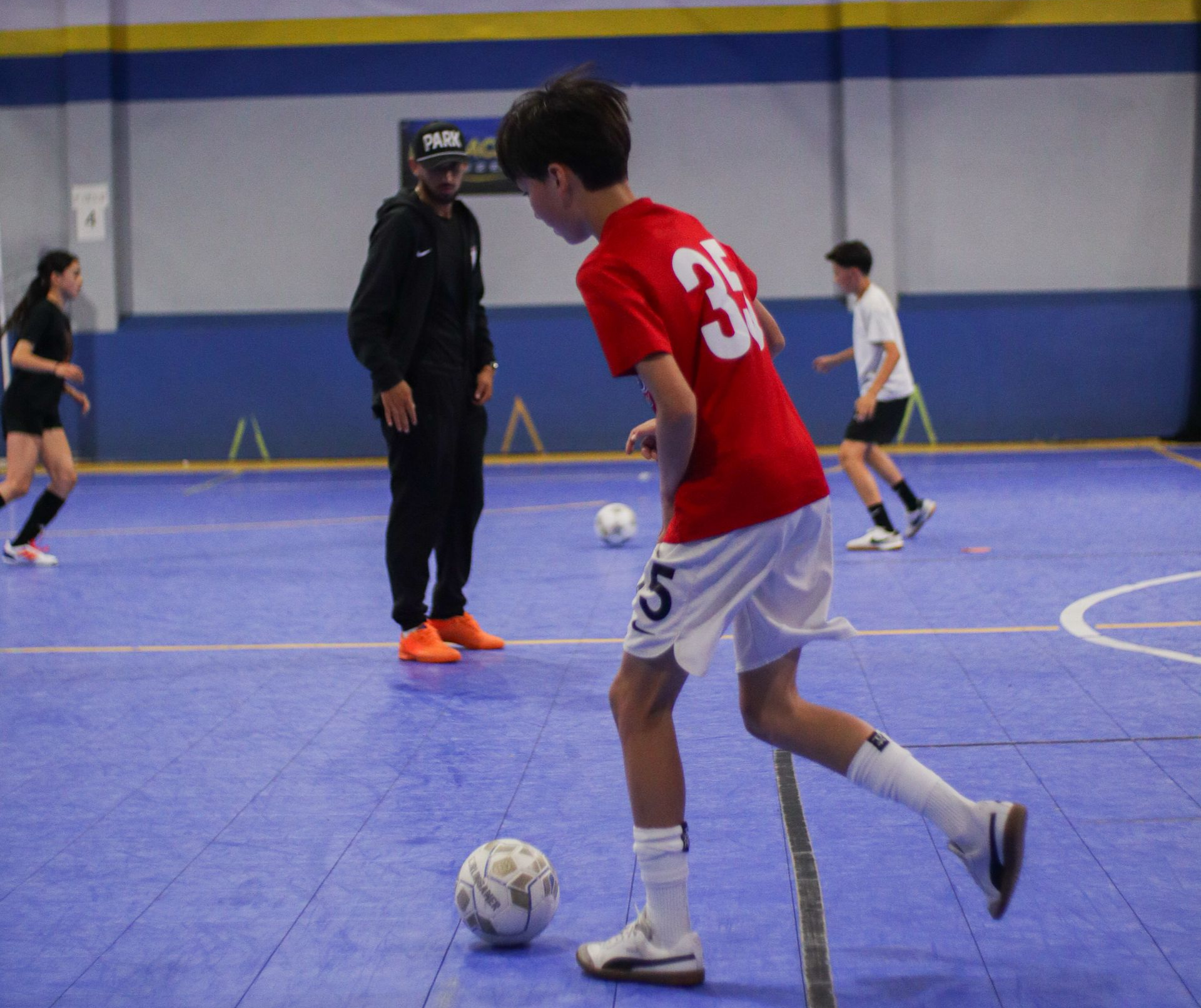 Boy in red soccer uniform practicing with ball indoors, coach watching. Bay city Futsal