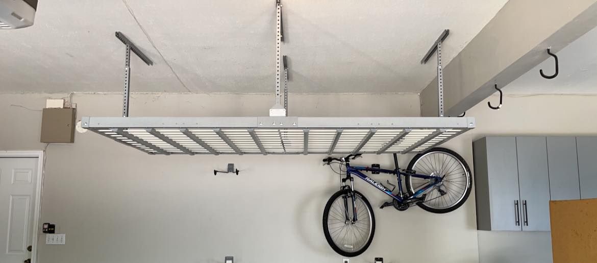 A bicycle hangs below a ceiling-mounted storage rack in a garage.
