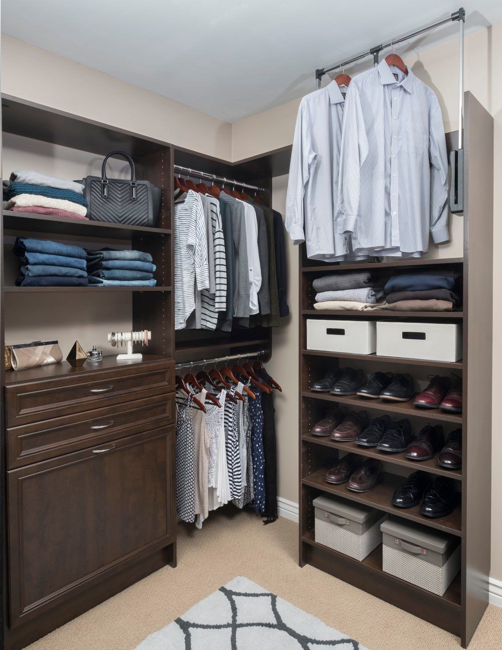 Walk-in closet with brown shelving, clothes, shoes, and folded items. Beige walls, carpet, and a rug.