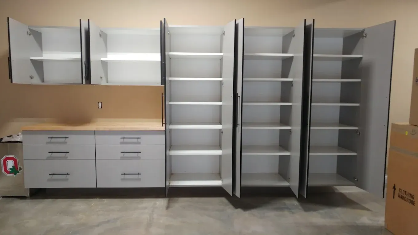 Garage cabinets: white with light wood countertop, open doors revealing shelves, against tan and beige walls.