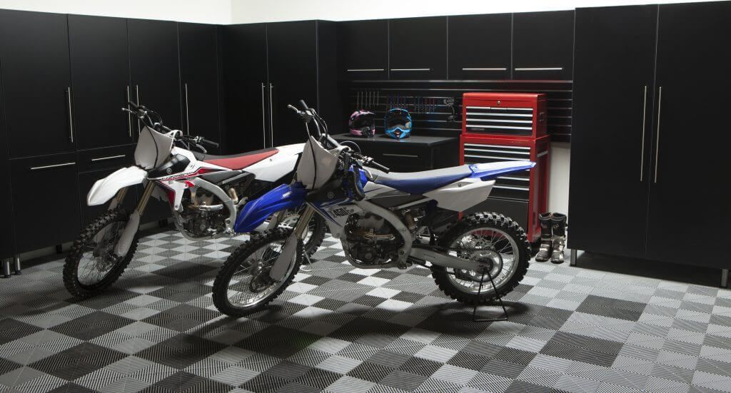 Two dirt bikes parked in a garage with black cabinets and checkered flooring.