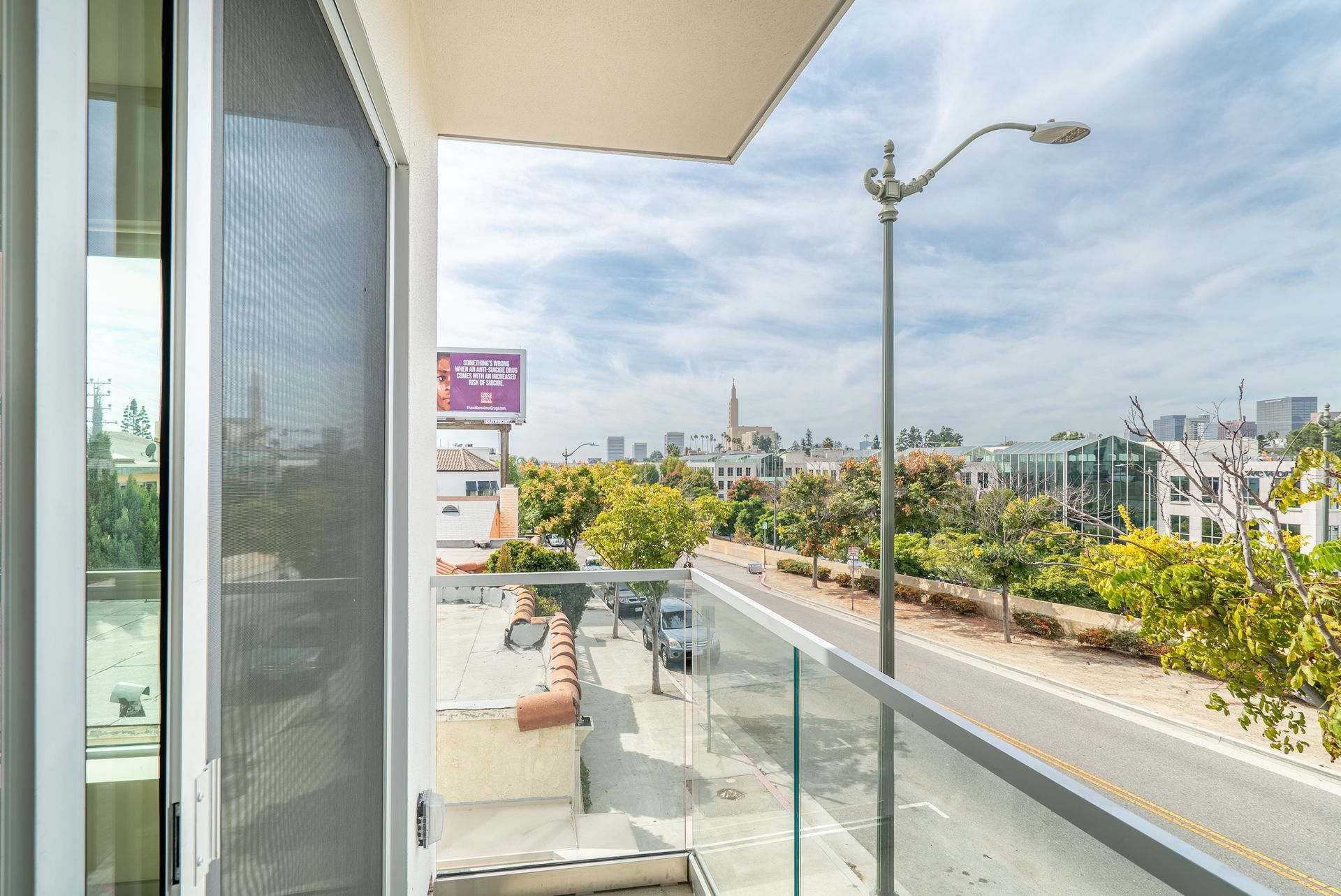 Balcony view overlooking a city street and skyline. Glass railing, overcast sky, trees.