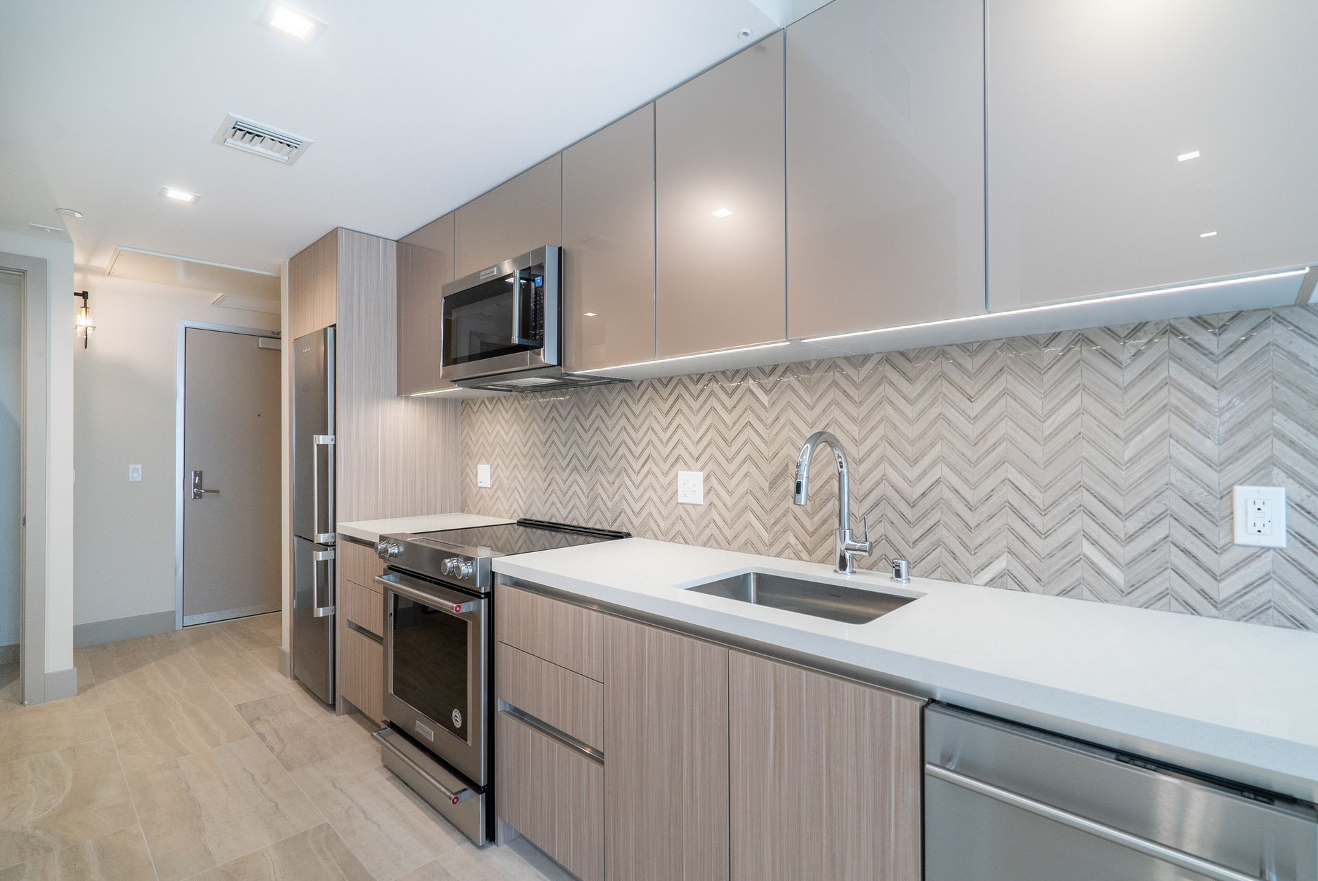 Modern kitchen with stainless steel appliances, light wood cabinets, and gray herringbone backsplash.