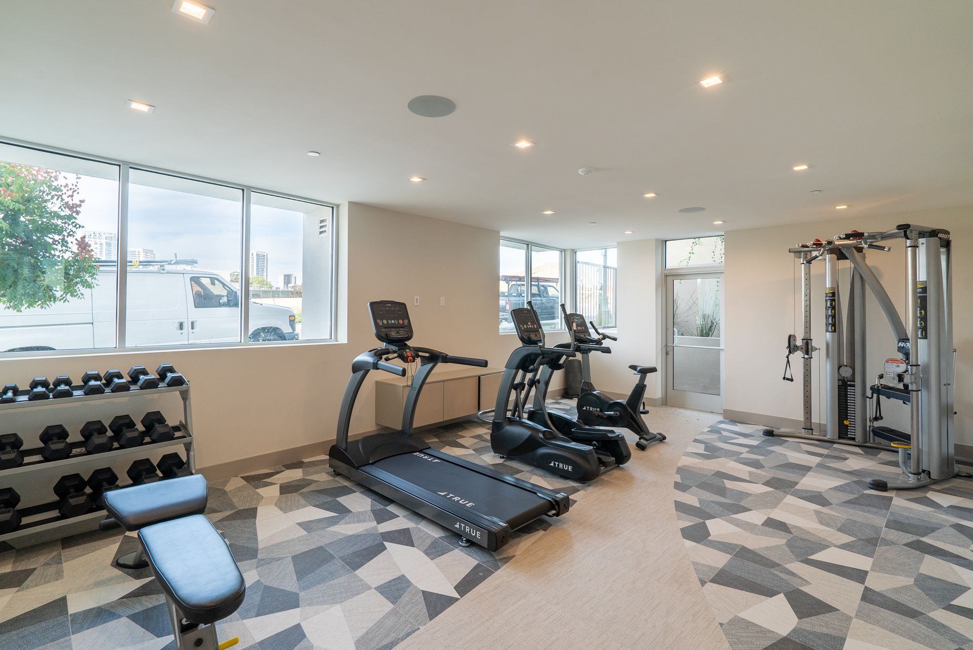 Gym with dumbbells, treadmills, stationary bike, weight machine, and a bench; windows along one wall.