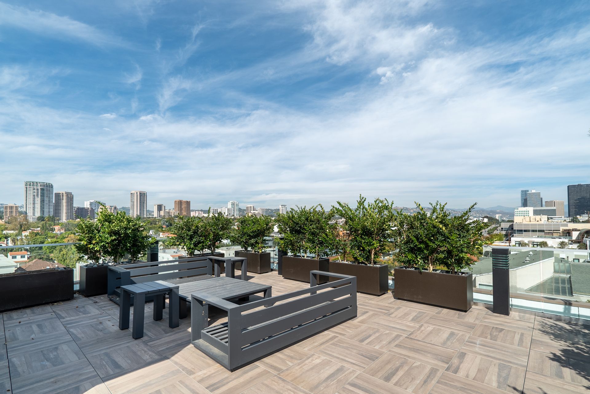 Rooftop patio with gray furniture, potted green plants, and city skyline under a blue, cloudy sky.
