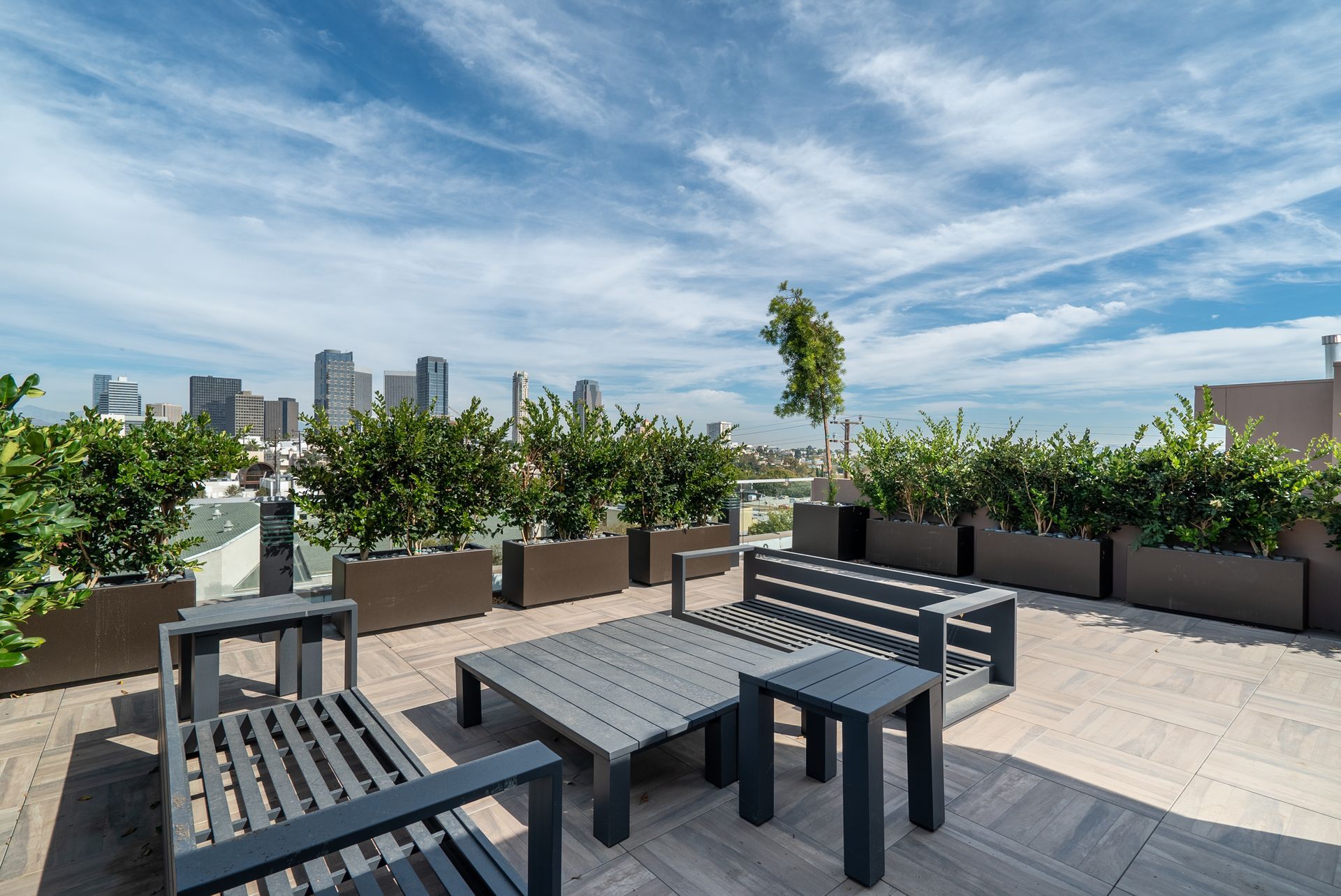 Rooftop patio with gray furniture, greenery, and city skyline under a blue sky with clouds.