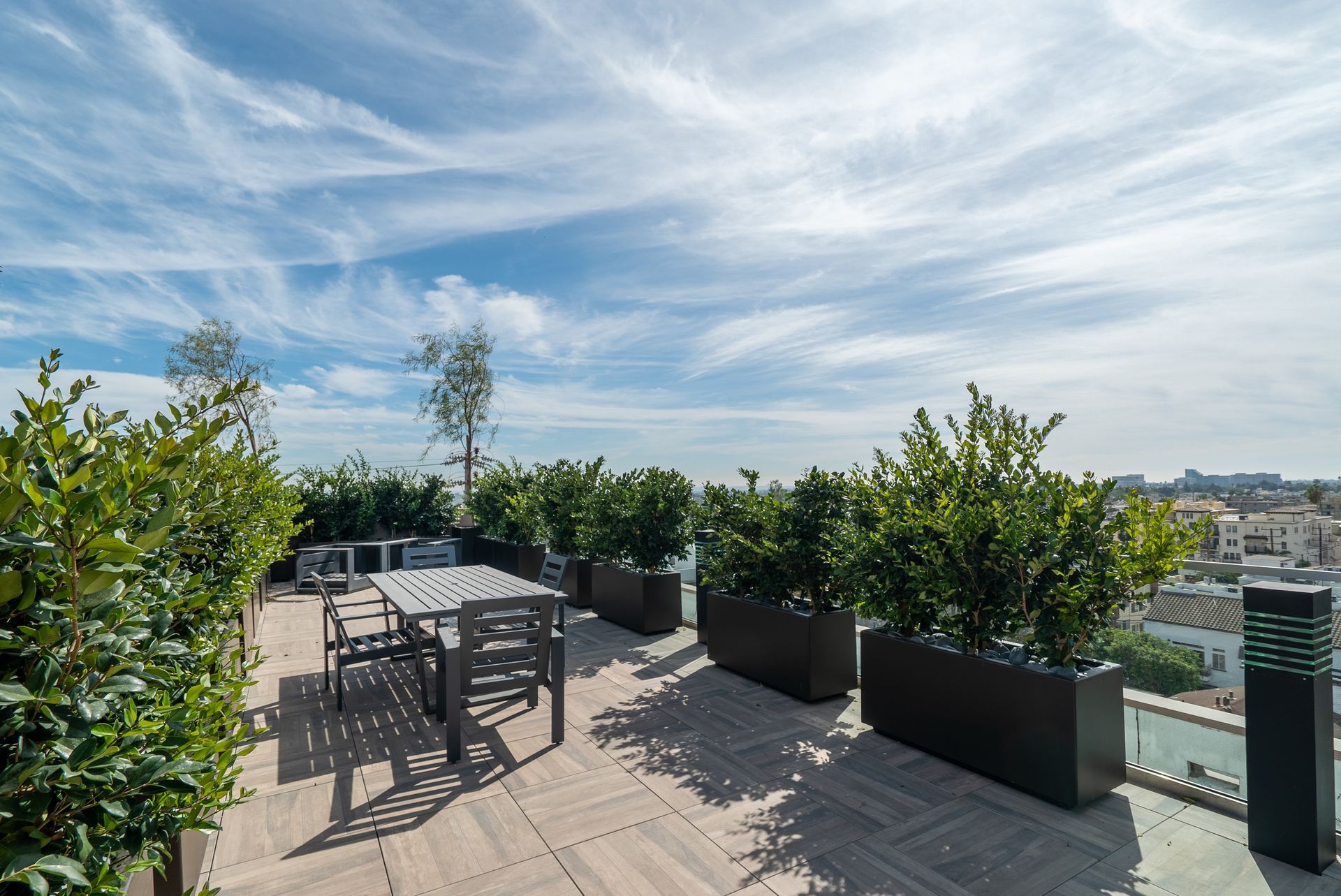 Rooftop patio with table, chairs, and potted greenery against a cloudy blue sky and city view.