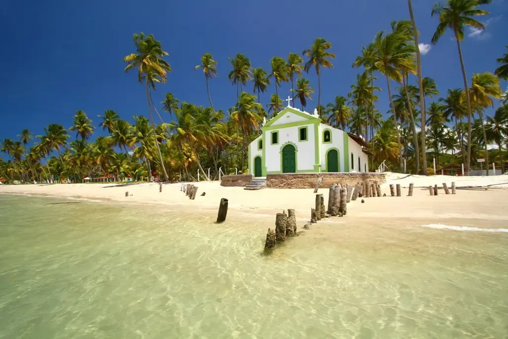 Uma igreja verde e branca está situada no topo de uma praia arenosa cercada por palmeiras.