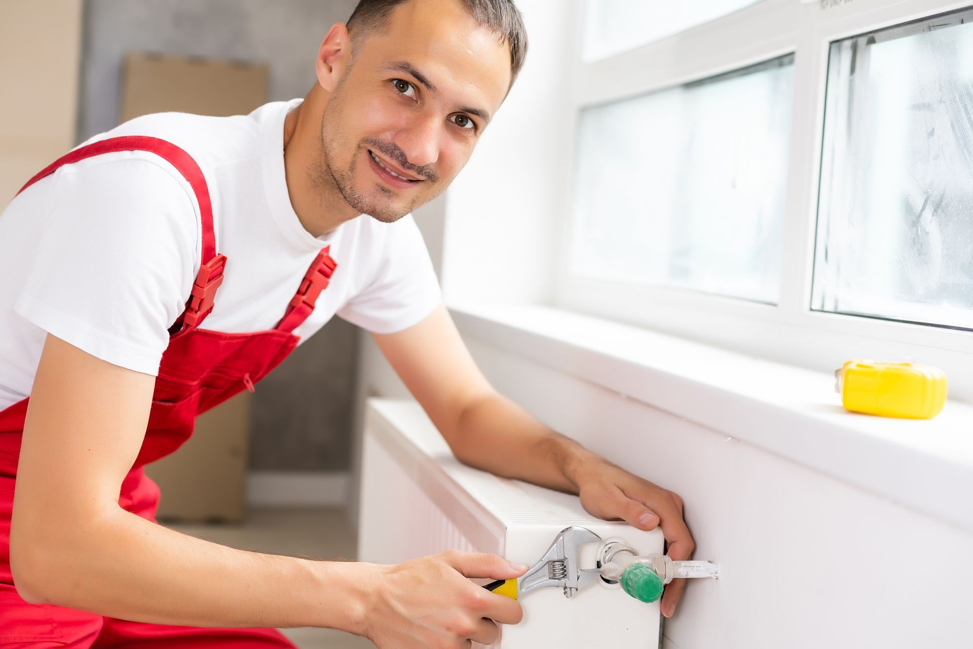 A heating contractor holding a wrench is installing a new heating system by a window.