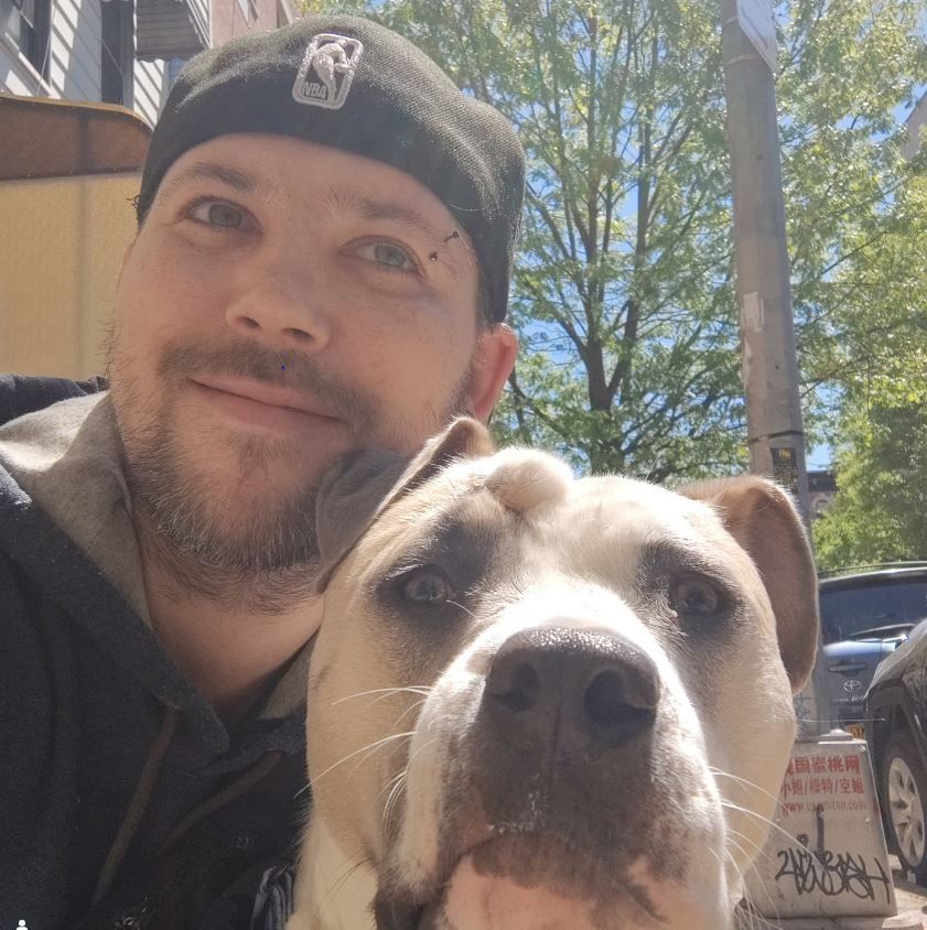 Man in black cap smiles, takes selfie with a large, light-colored dog outdoors on a sunny day.