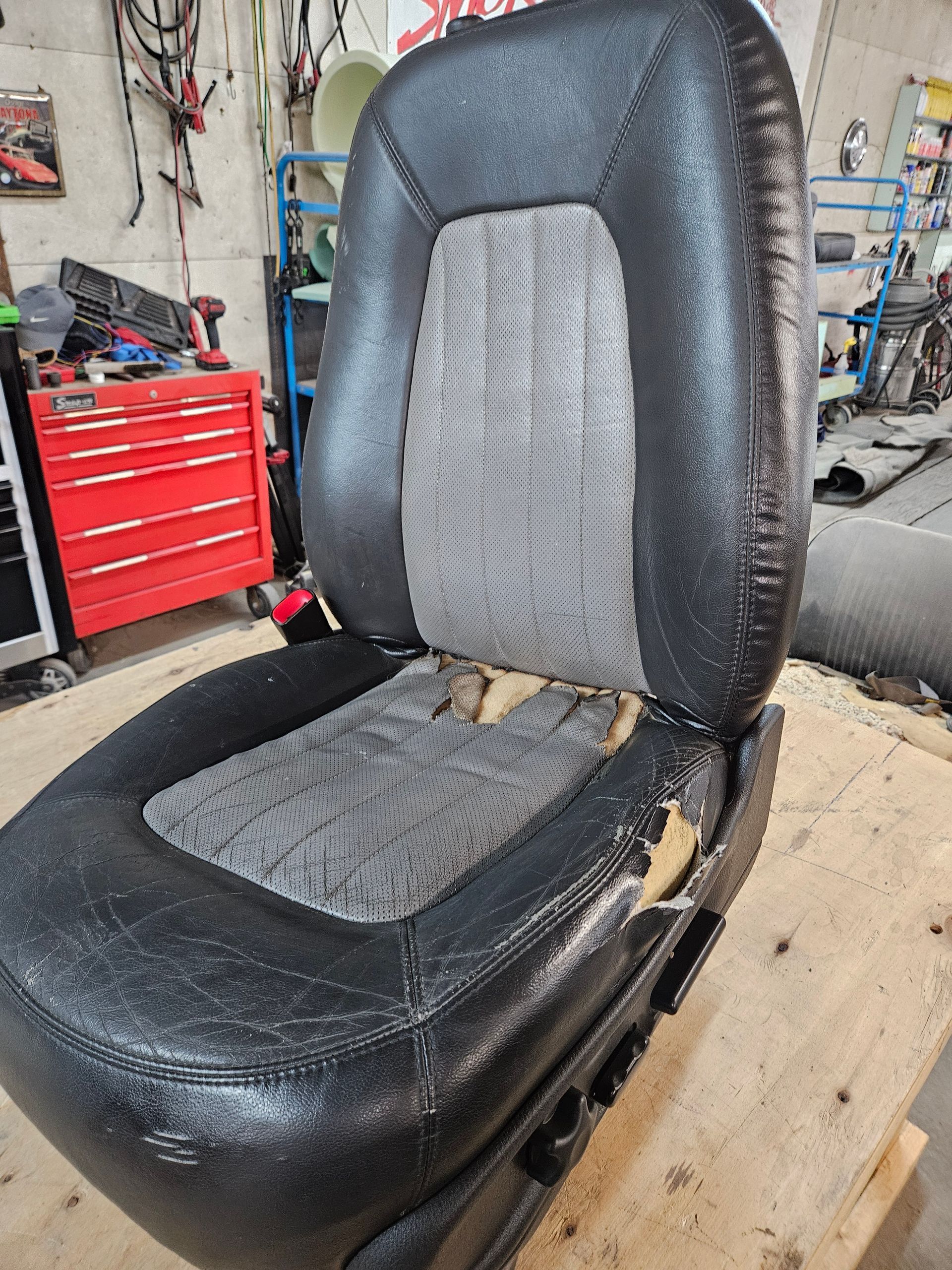 Black car seat with gray inserts on a workbench in a garage, with tools and a red toolbox nearby