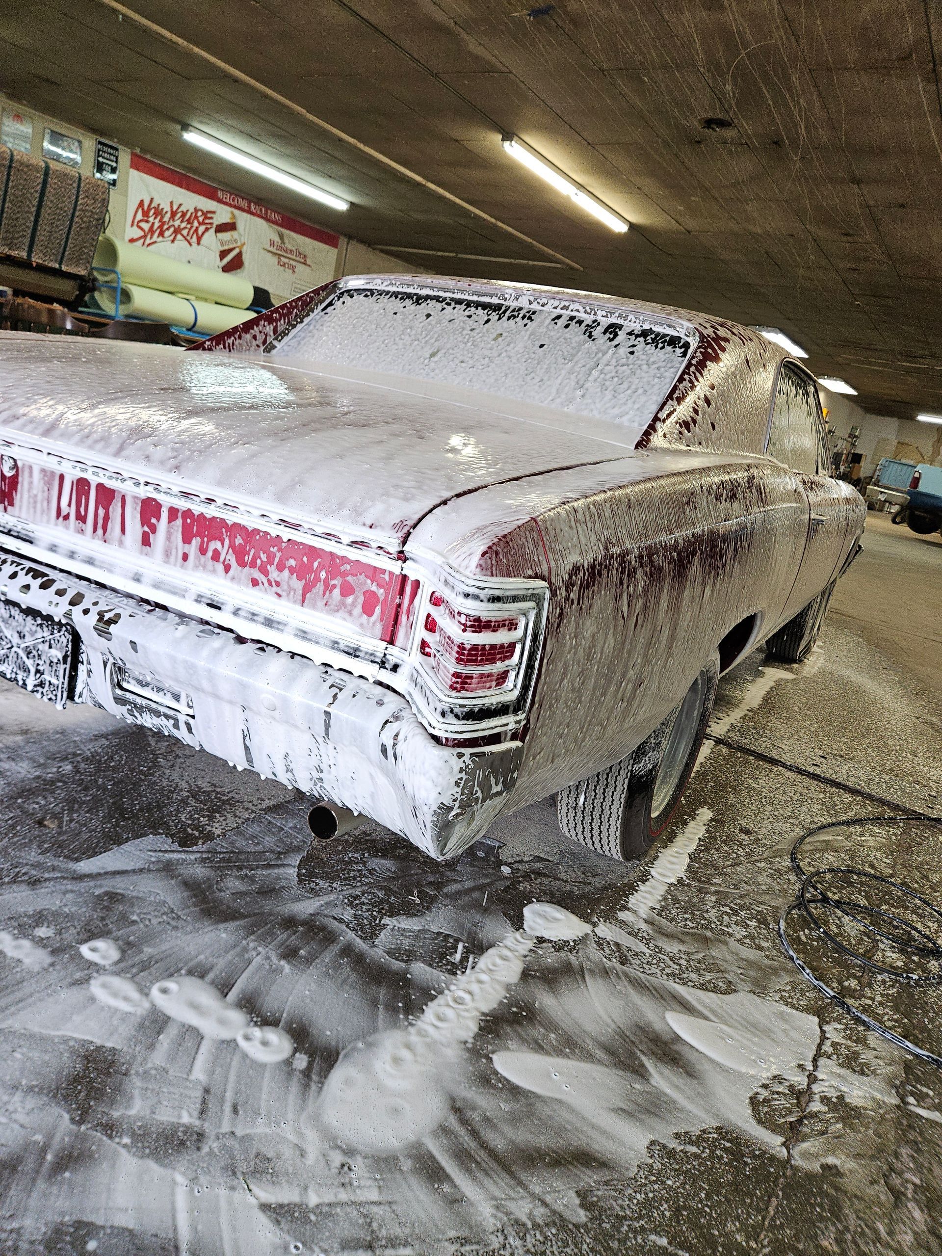 Foam-covered white car in an automatic car wash, with soap and water on the floor.
