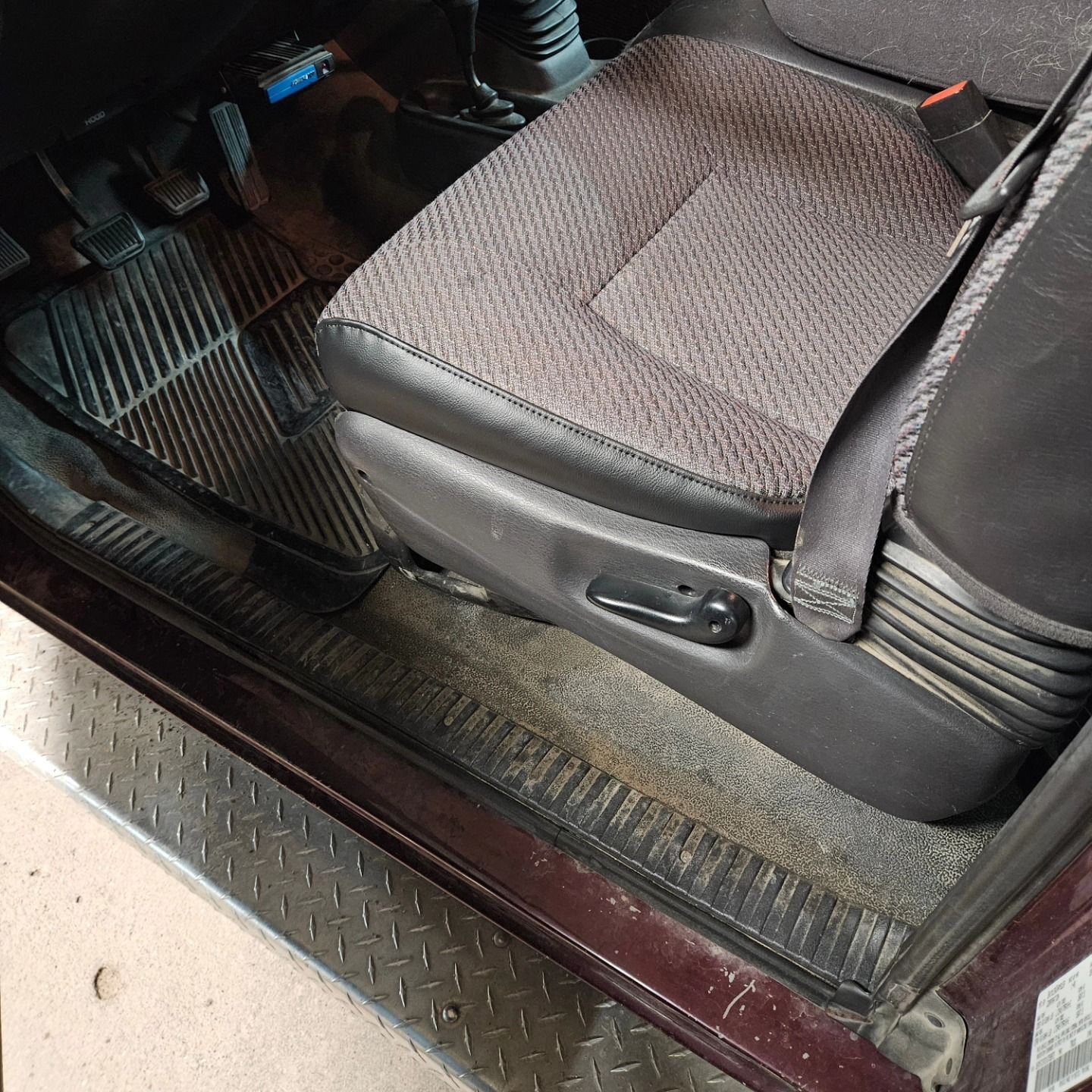 Car interior showing gray cloth front seat with manual adjustment lever and rubber floor mat