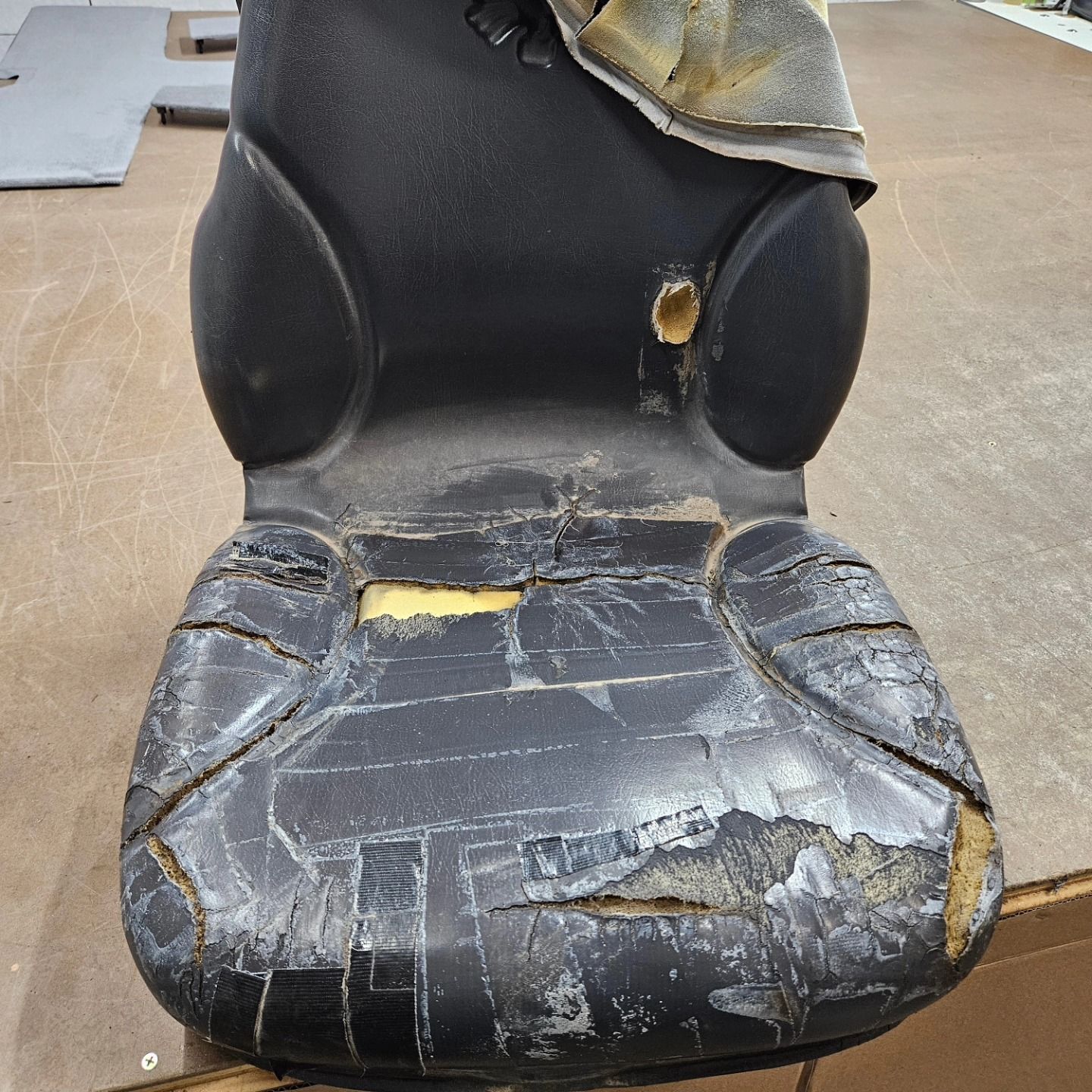 Worn black office chair with torn, peeling seat upholstery in a workshop or warehouse.