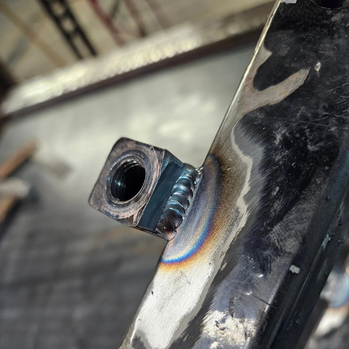 Close-up of a welded metal bracket with a threaded hole and rainbow heat discoloration on black steel.