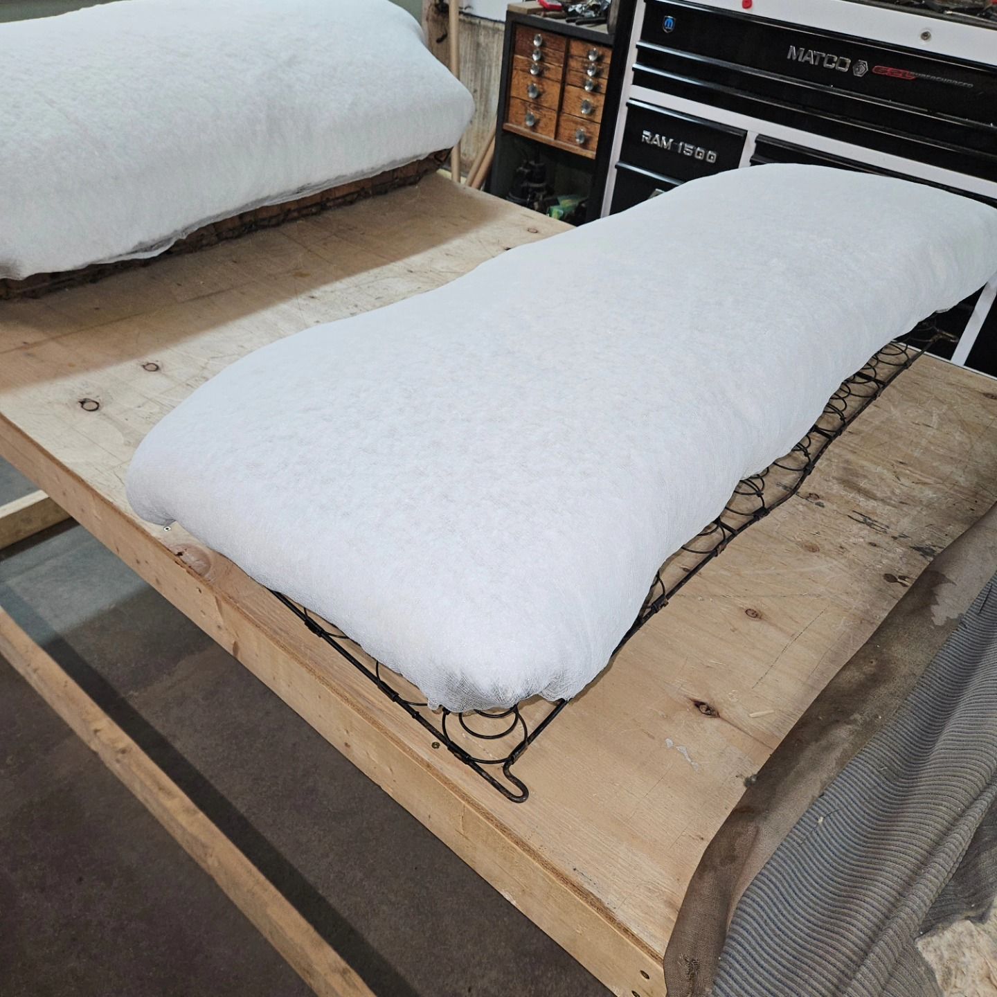 Two white foam cushions on a wooden bed frame in a workshop or furniture showroom.